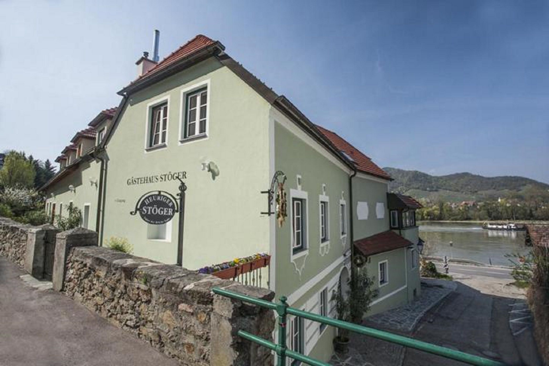 A "Gästehaus Stöger" és a "Hagyományos borozó Stöger" feliratú zöld vendégház egy folyó partján.