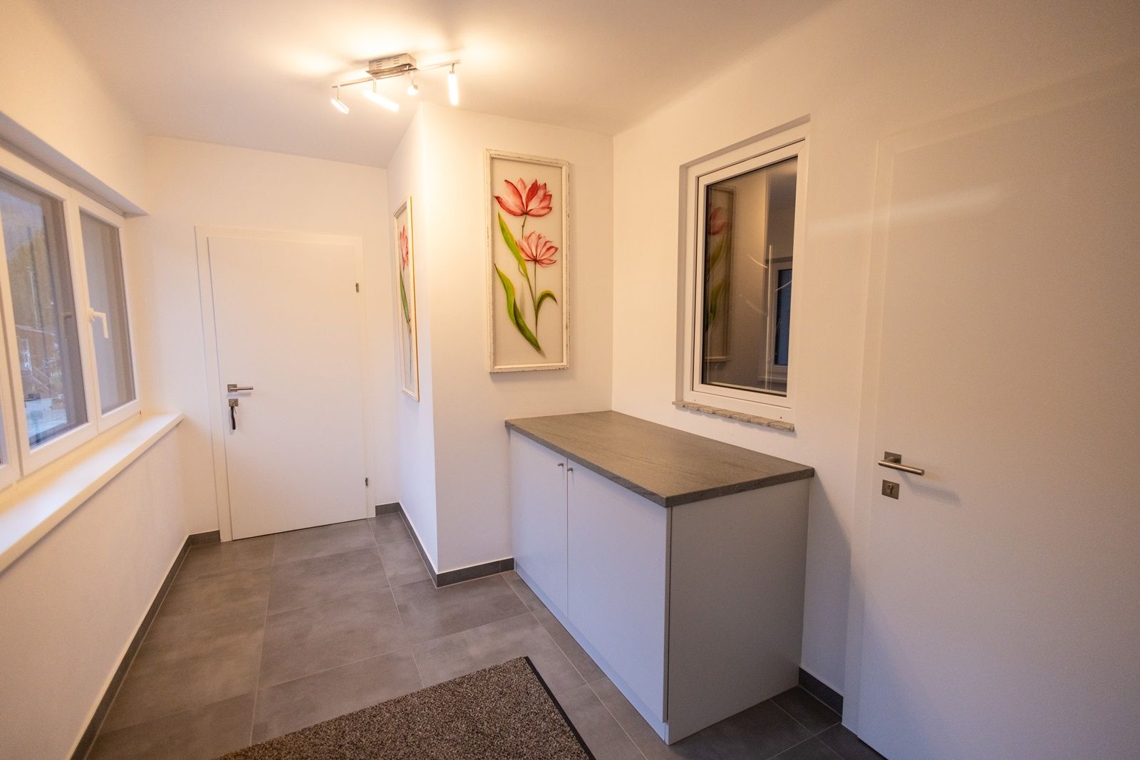 Modern előszoba fehér falakkal, szürke padlóval, szekrénnyel és virágos képekkel a falon.