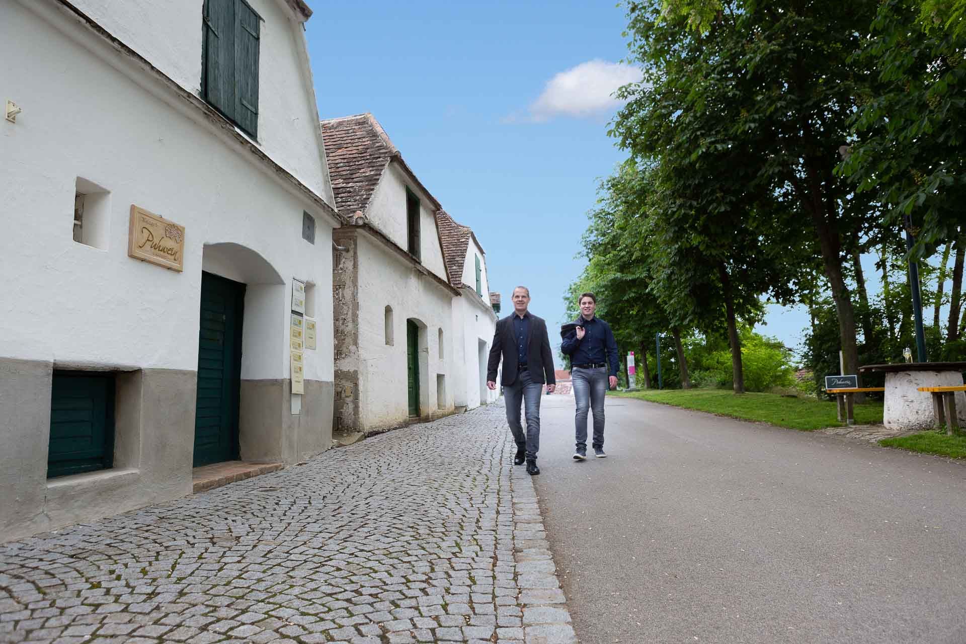 Két férfi sétál egy macskaköves utcán, fehér épületek és fák mellett.