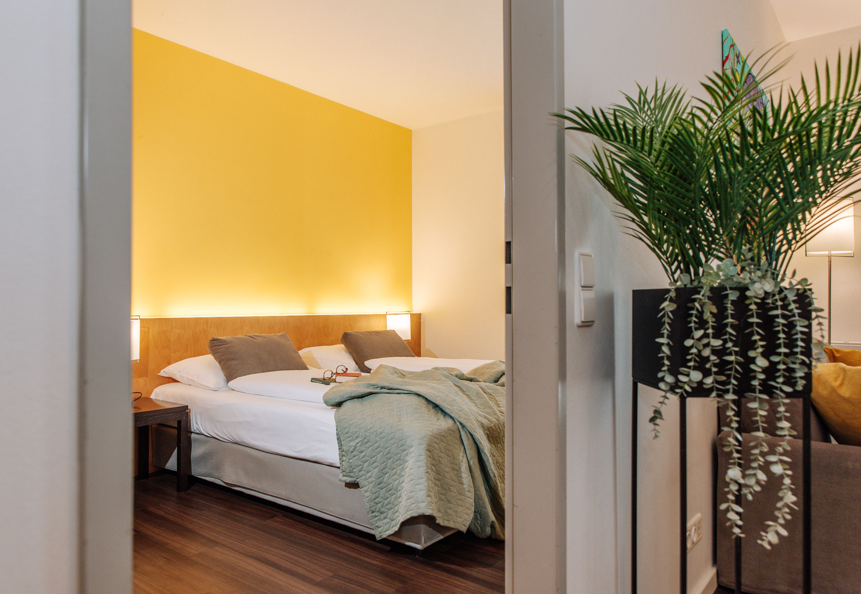 Szállodai szoba sárga fallal, franciaággyal és növénnyel az előtérben.