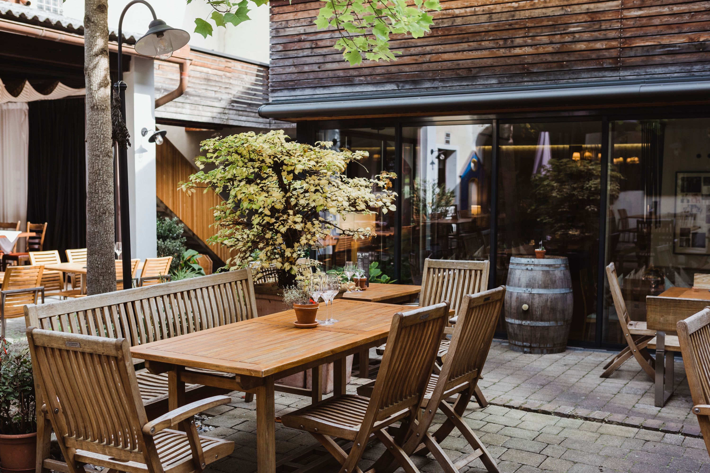 Hangulatos udvar fából készült bútorokkal, növényekkel és boroshordóval, amelyet fából készült burkolatokkal díszített épületek vesznek körül.