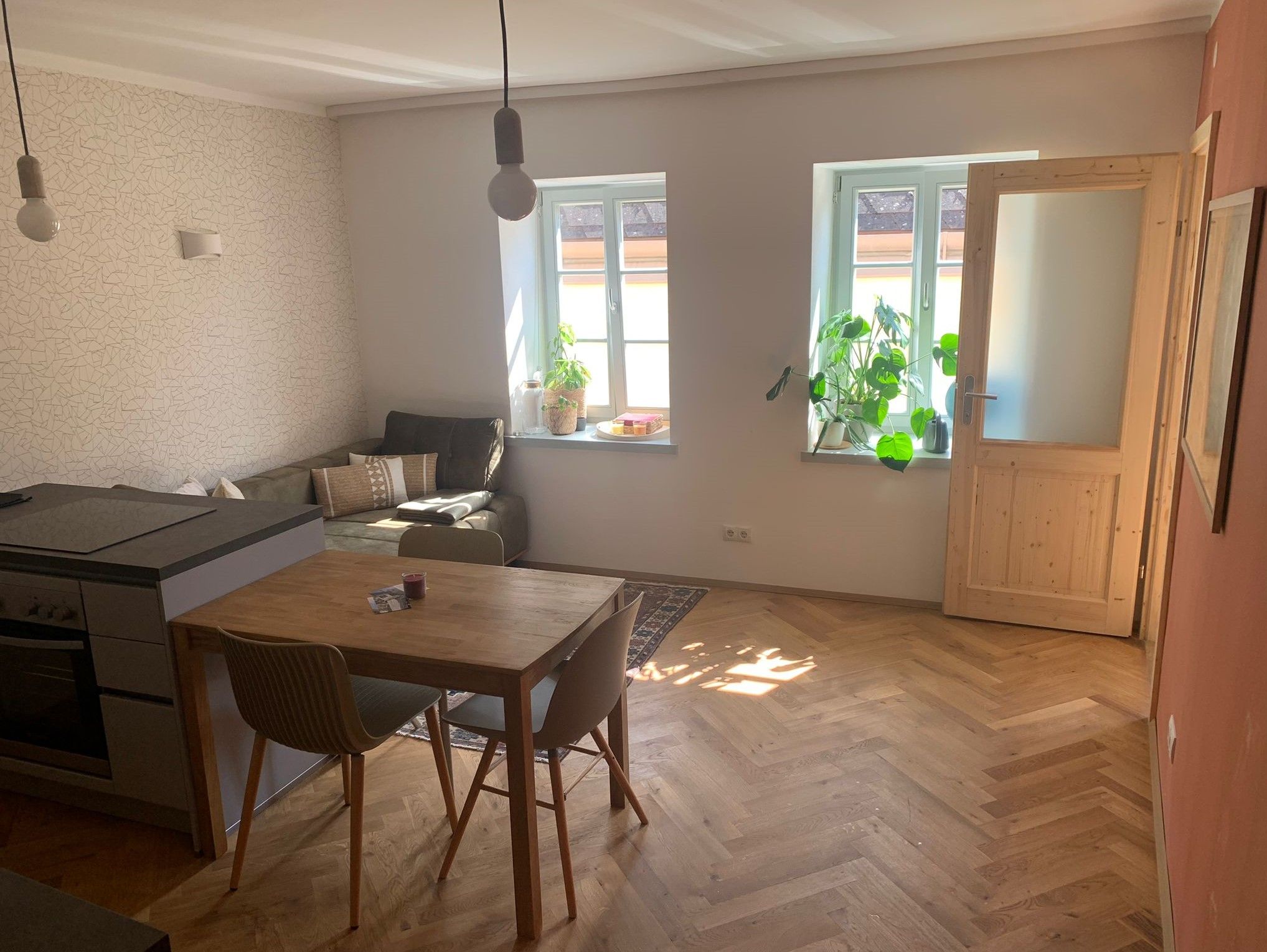 Világos nappali fapadlóval, étkezőasztallal, kanapéval és növényekkel az ablak mellett.