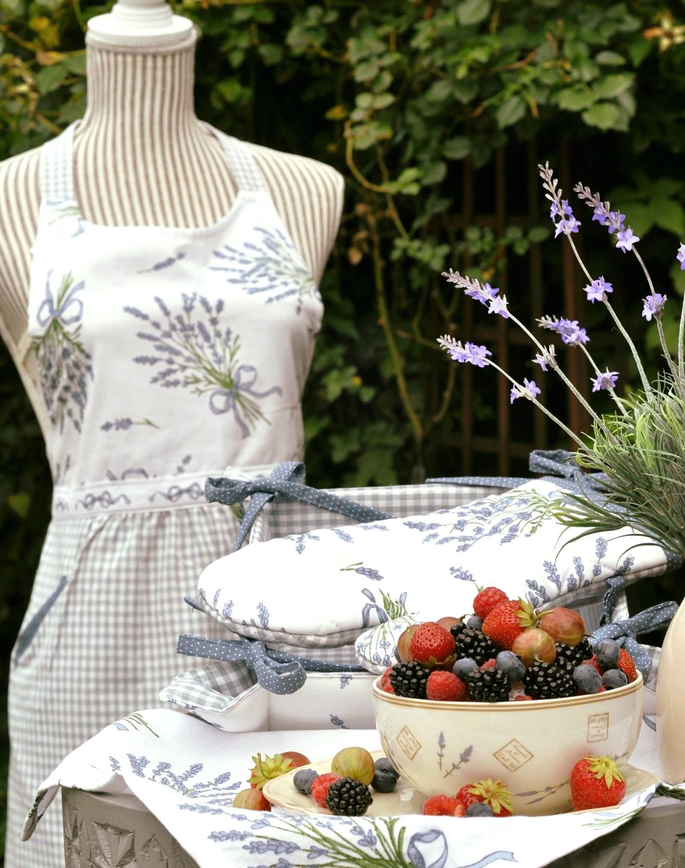 Egy tál bogyós gyümölcsökkel egy asztalon, levenduladíszítéssel és egy kötényes szabóbábuval a háttérben.