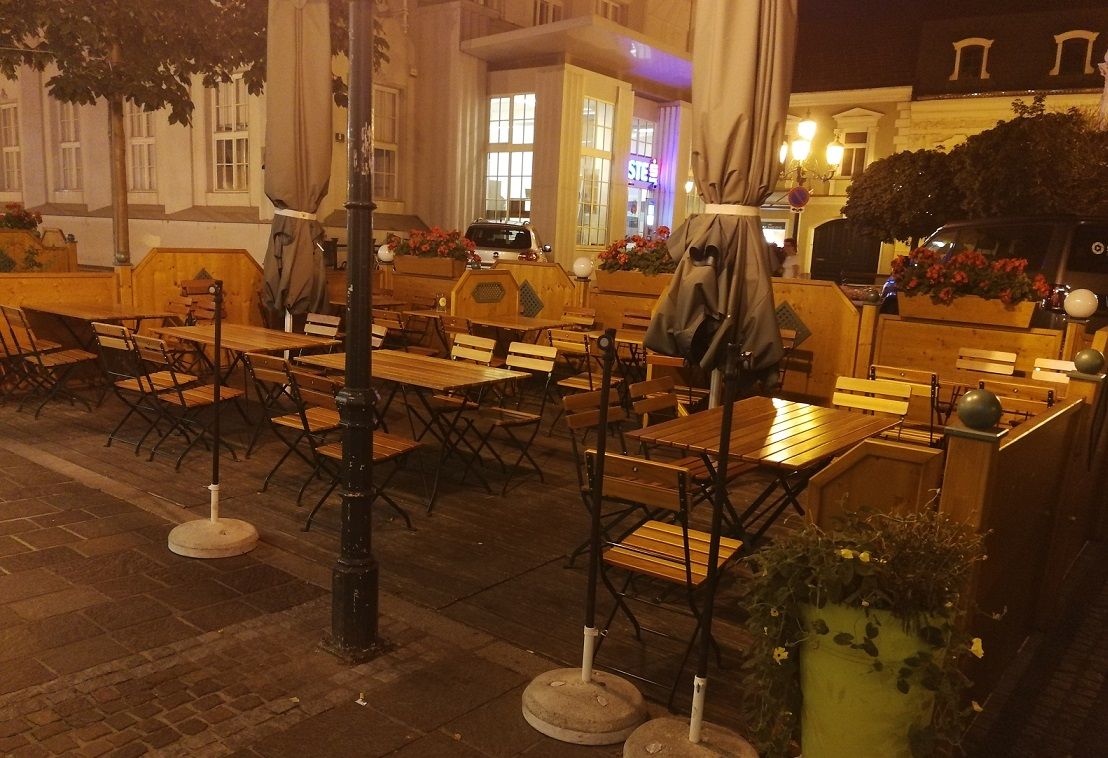 Egy étterem szabadtéri területe üres faasztalokkal és székekkel, növényekkel és lámpásokkal körülvéve éjszaka.