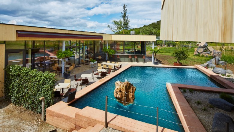 Modern terasz asztalokkal és székekkel egy téglalap alakú tavacska mellett, amelynek közepén sziklák vannak.