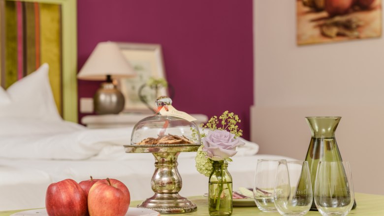 St&iacute;lusosan berendezett szoba, egy asztallal, amelyen alma, keksz egy &uuml;vegharangl&aacute;b alatt, egy v&aacute;za vir&aacute;ggal &eacute;s poharak vannak elhelyezve.