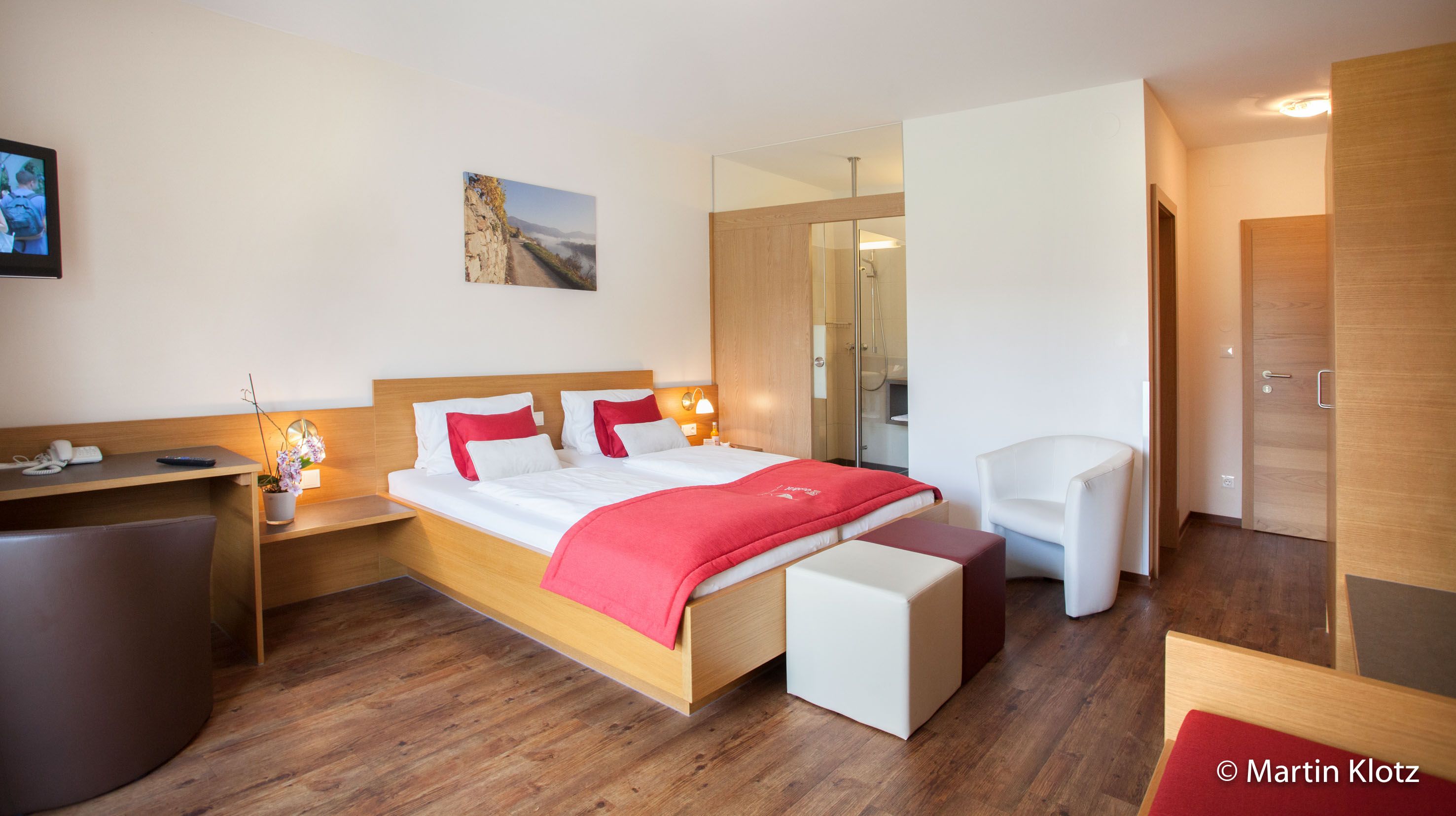 Modern szállodai szoba franciaággyal, piros díszítéssel, íróasztallal és TV-vel.