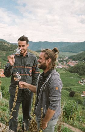 Inmitten der sanften Hügel und Weinreben genießen zwei Freunde den Augenblick mit einem Glas Wein in der Hand. Die malerische Landschaft der Donau in Niederösterreich bietet eine perfekte Kulisse für entspannte Stunden in der Natur.