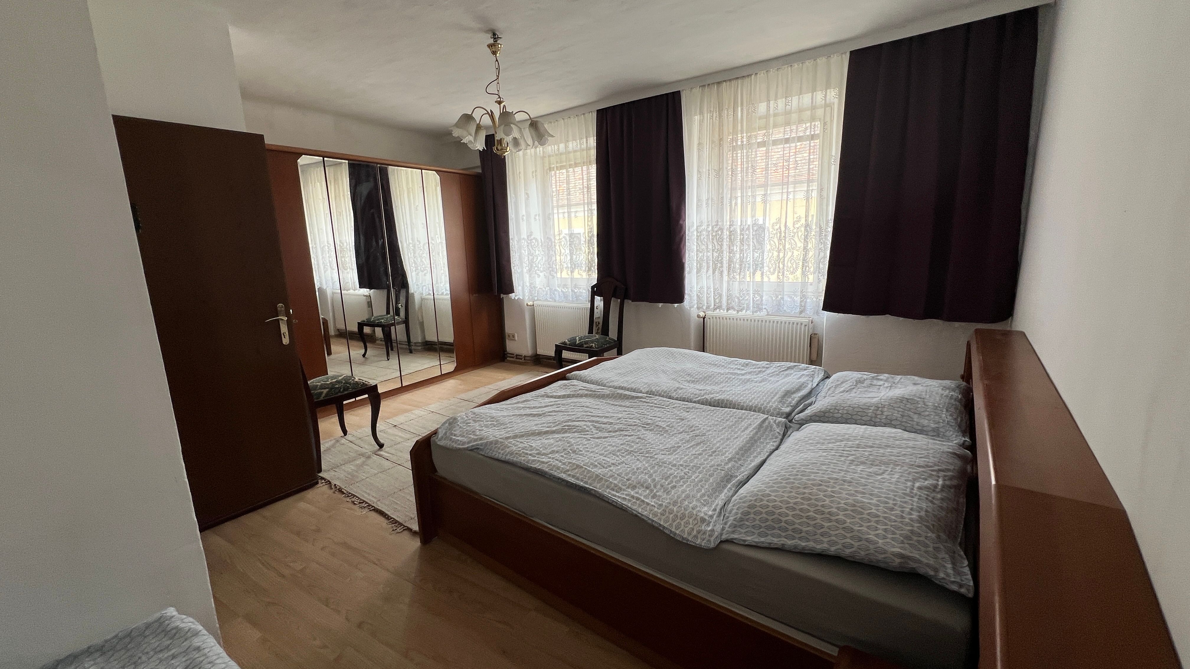 Hálószoba franciaággyal, tükrös szekrénnyel, székkel és függönyökkel az ablakokon.