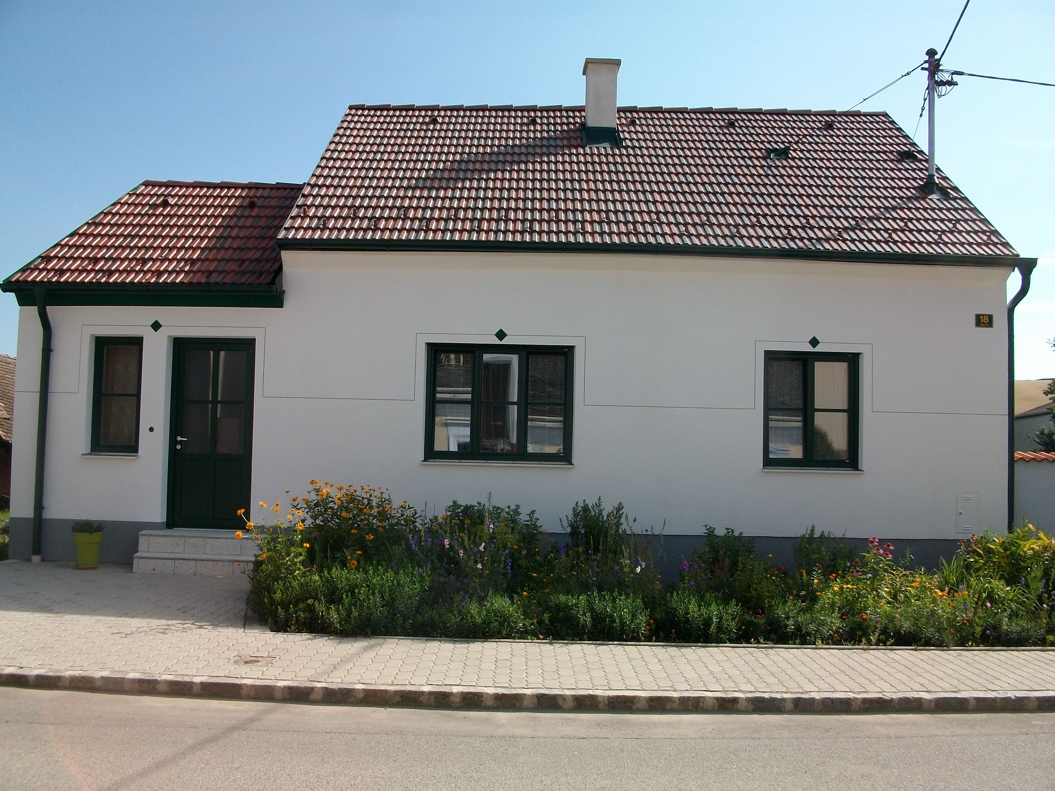 Kis, fehér ház, piros cseréptetővel és zöld ajtókerettel, körülötte gondozott, virágos kert.