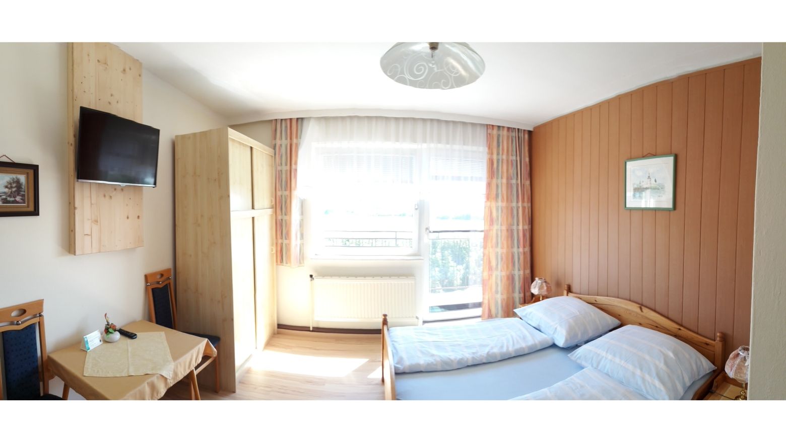 Hangulatos vendégszoba franciaággyal, fabútorokkal, kis asztallal és a falon található TV-vel.