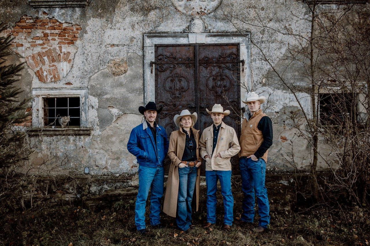 Négy cowboy-öltözetű ember áll egy régi, romos fal előtt, amelyen egy nagy fémajtó van.