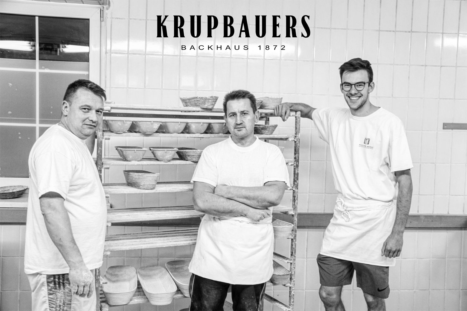 Három pék egy pékségben, a háttérben kenyérkosarakkal.