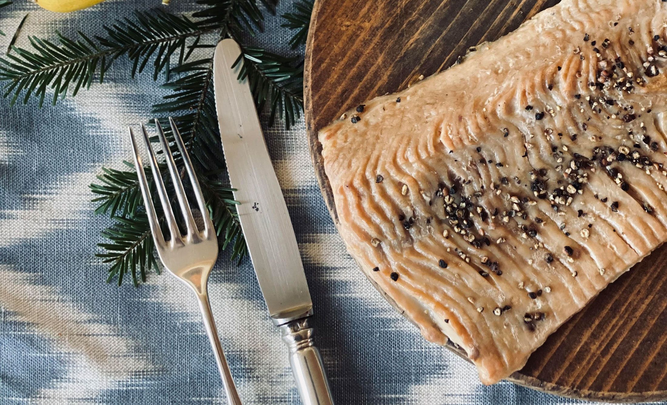 Közelkép egy füstölt pontyfiléről fatányéron, borsszemekkel díszítve. Mellette egy villa és egy kés egy kék ruhán.