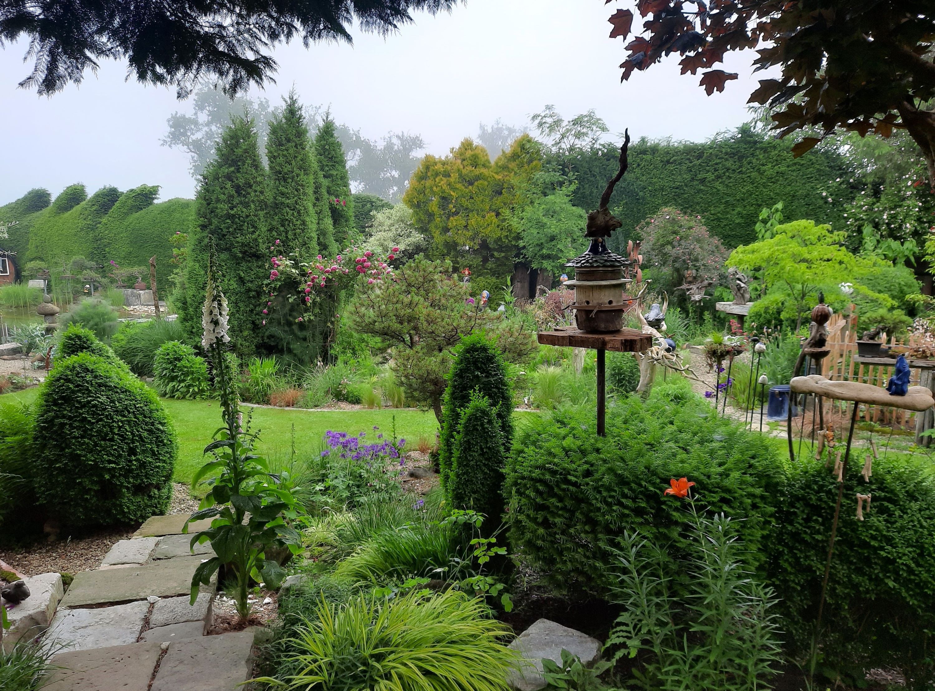 Egy buja kert különböző növényekkel, fákkal és dekorációkkal. Egy madárház áll a középpontban.