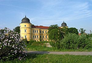 Sárga kastély két toronnyal és piros tetővel, fákkal és virágokkal körülvéve.
