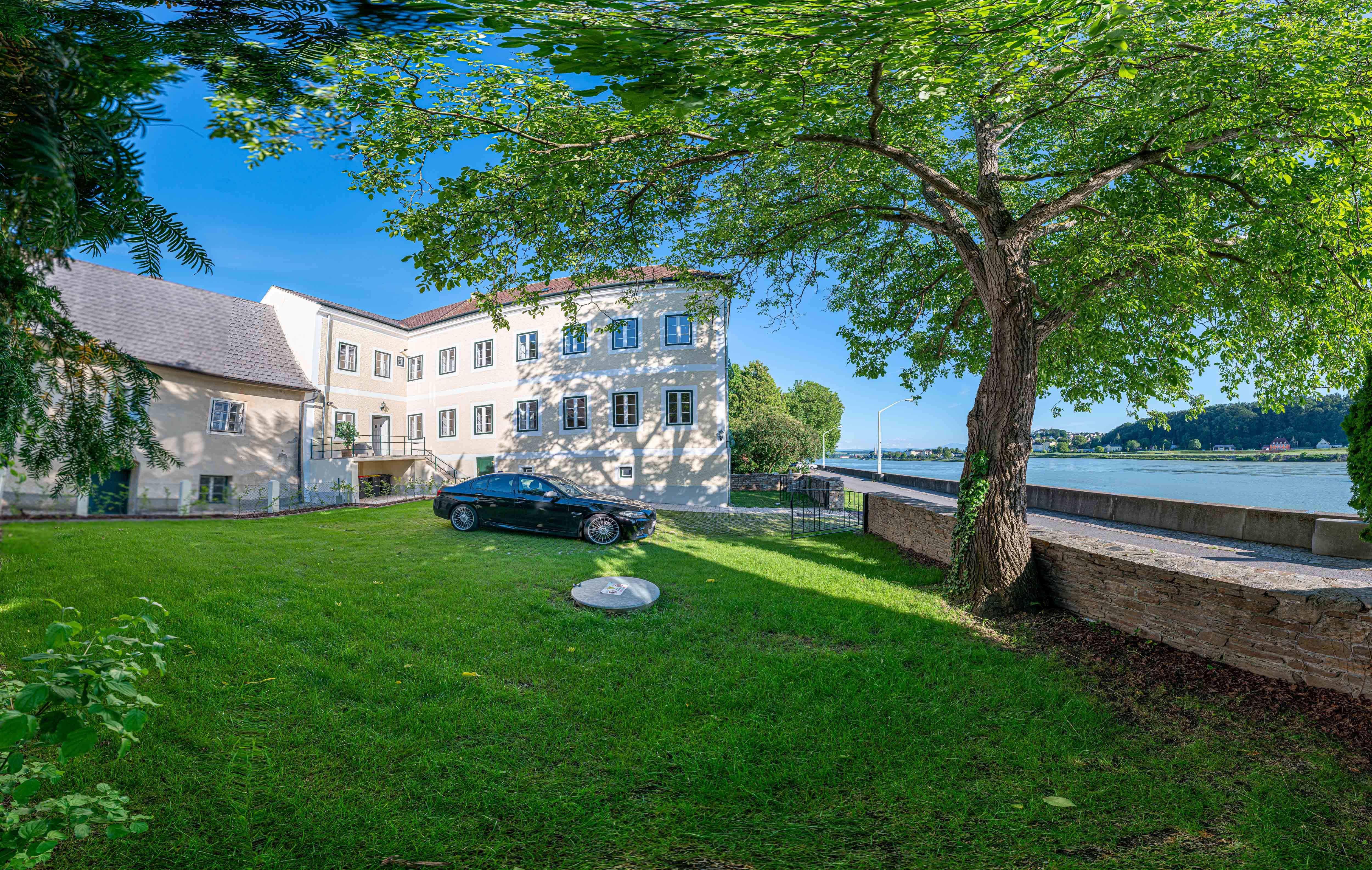 Egy régi posta külső nézete, előtérben fa és autó, háttérben a Duna.