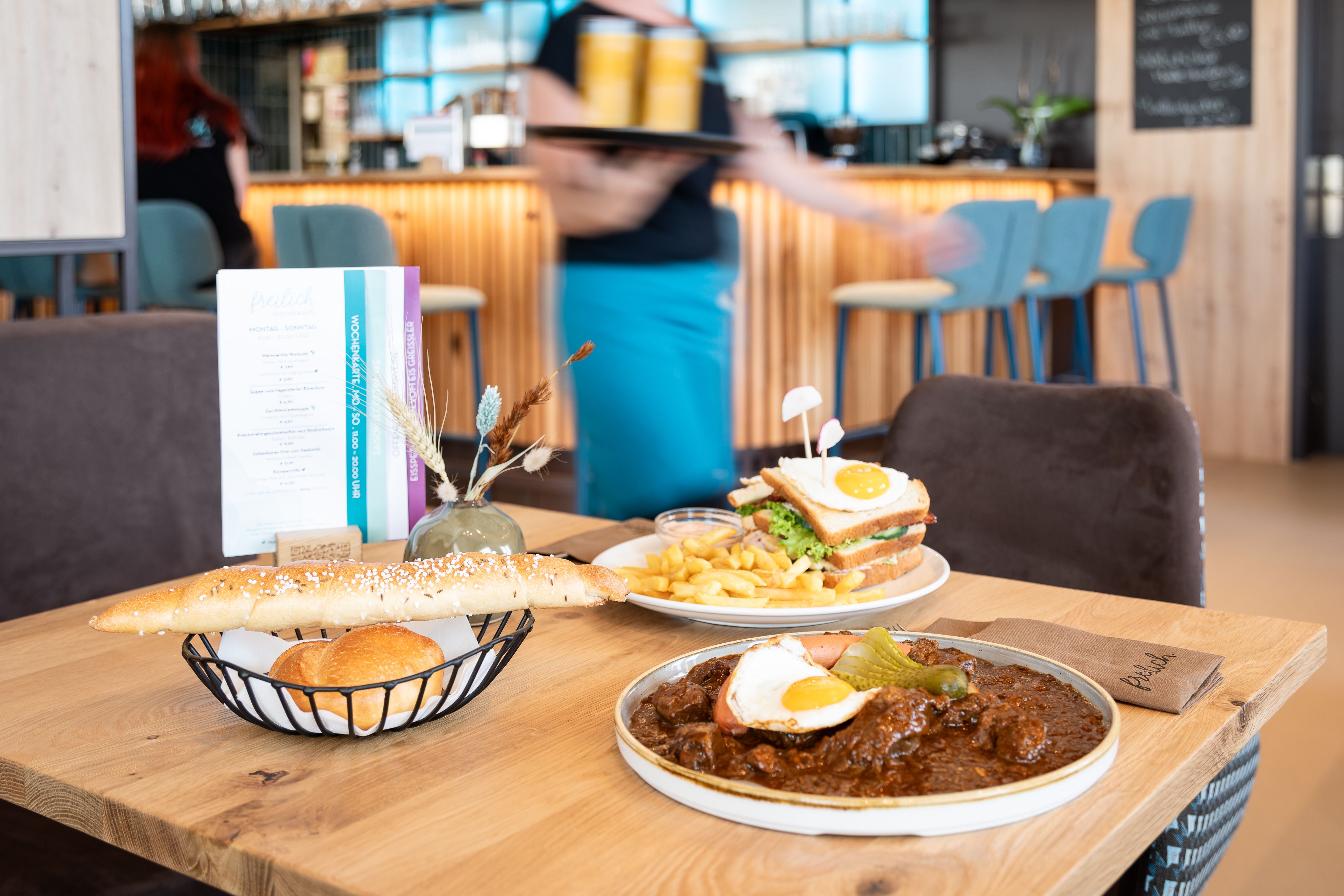 Egy asztal egy étteremben, regionális ételekkel: egy tányér gulyás és tükörtojás, egy szendvics chipsszel és egy kenyérkosár.