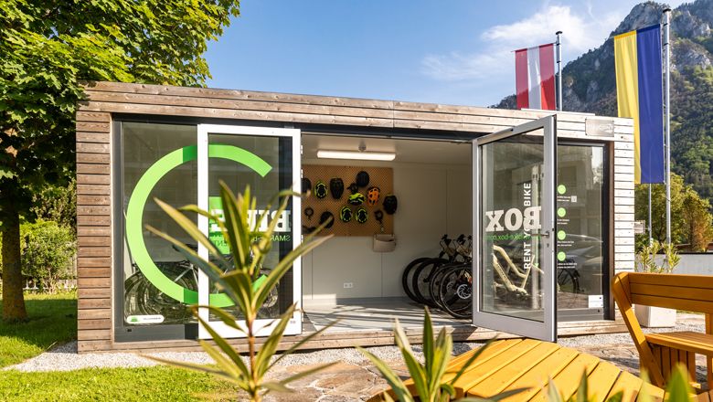 Egy modern kerékpáros doboz nyitott ajtókkal, kerékpárokkal és bukósisakokkal, természet és zászlókkal körülvéve.