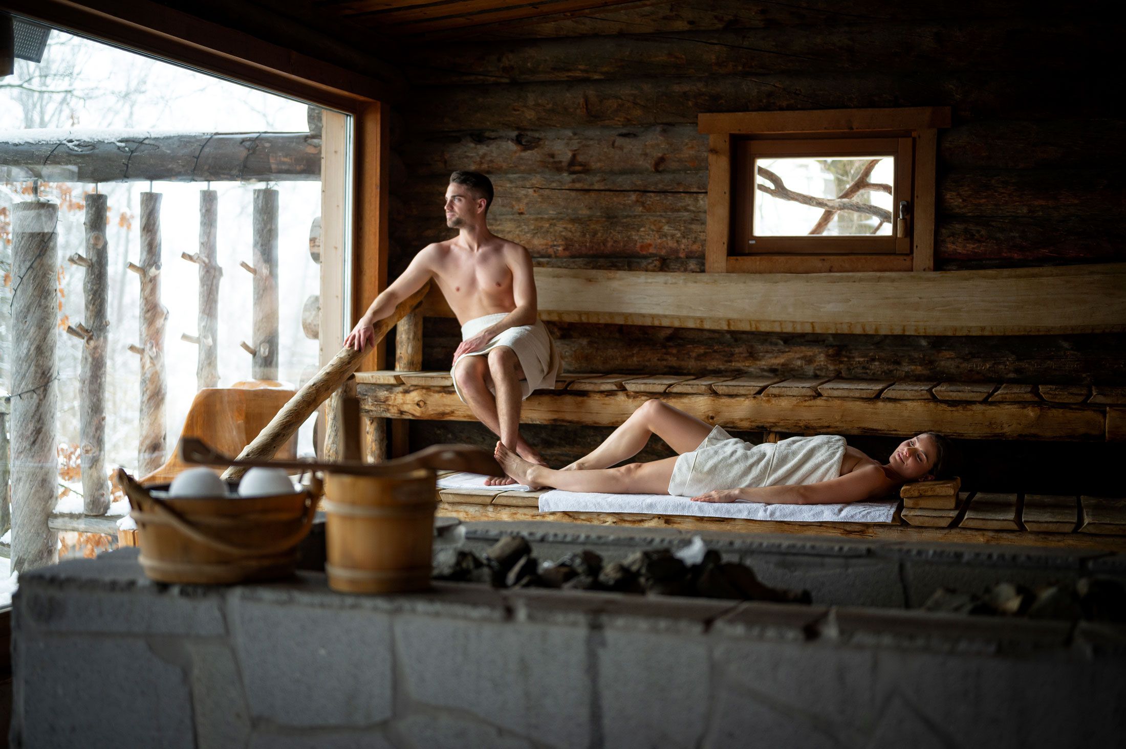 Két ember egy fából készült szaunában, az egyik ül, a másik fekszik, mindketten törölközőbe burkolózva.
