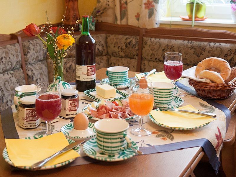 Megterített reggelizőasztal zsemlével, lekvárral, tojással, gyümölcslével és virágokkal.