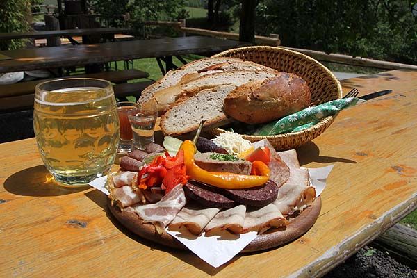 Deszkás uzsonna kenyérrel, kolbásszal, szalonnával és italokkal egy kinti faasztalon.