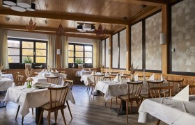 Elegáns étterem, fából készült bútorokkal, fehér terítőkkel és nagy ablakokkal, amelyeken őszi fák láthatók.