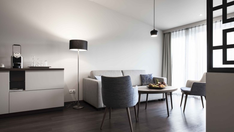 Modern &uuml;lősarok kanap&eacute;val, karossz&eacute;kkel, asztallal &eacute;s &aacute;ll&oacute;l&aacute;mp&aacute;val egy vil&aacute;gos szob&aacute;ban.