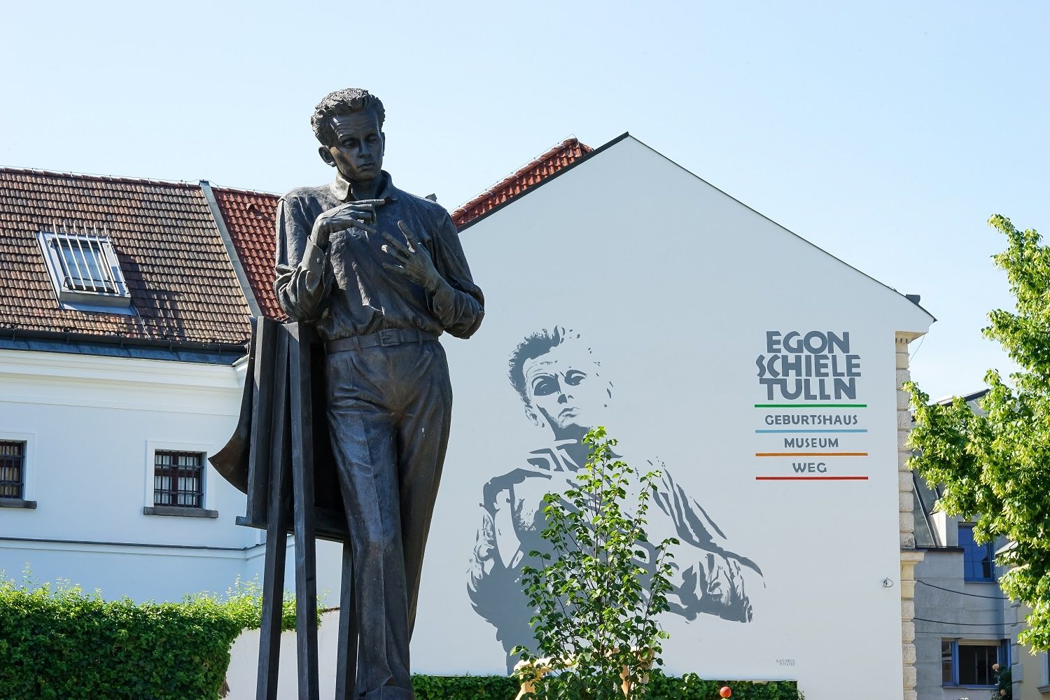 Egon Schiele szobra egy épület előtt, falfestéssel és "Egon Schiele Tulln" felirattal.