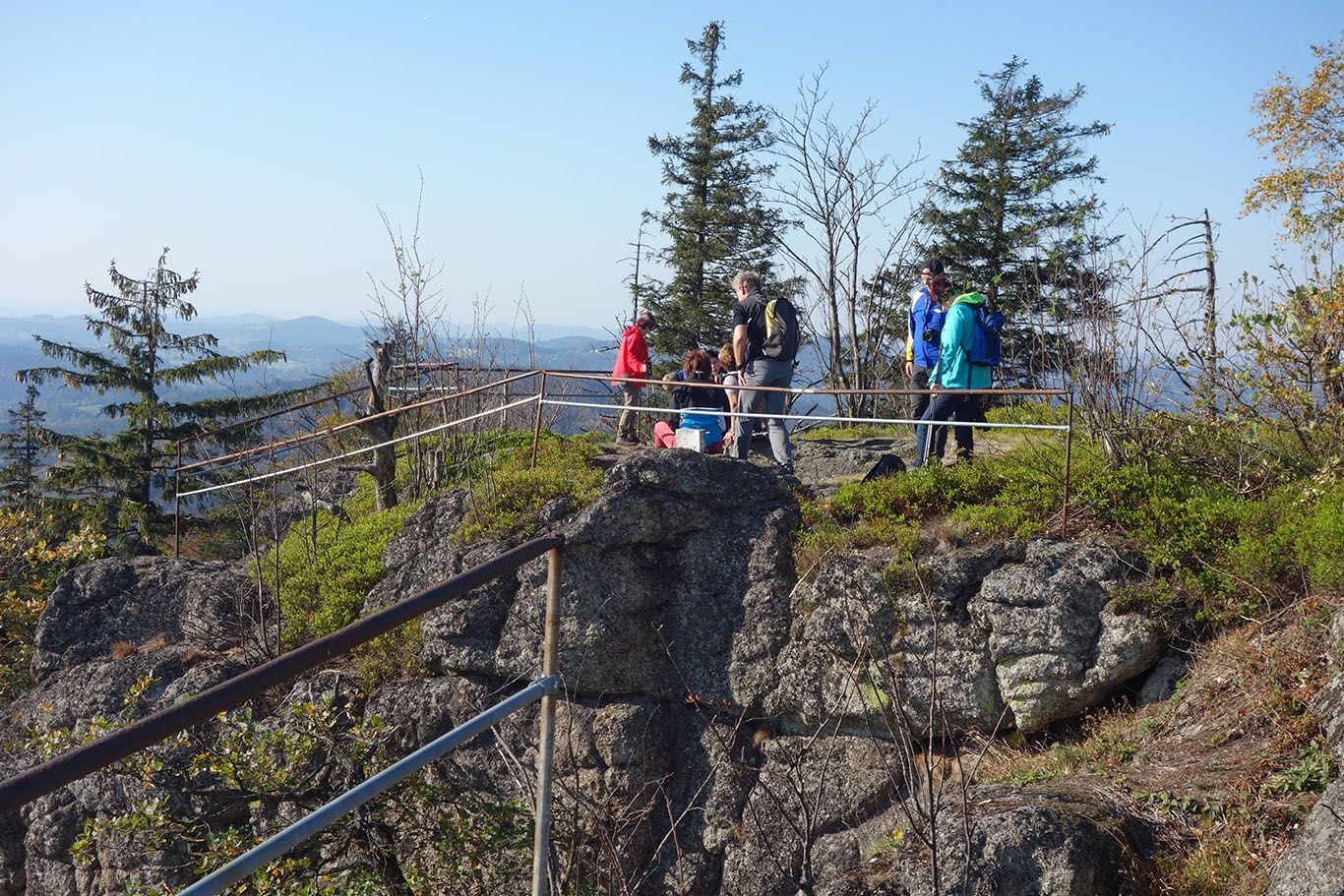 Túrázók csoportja egy sziklás, korlátos kilátóponton, a háttérben fák és hegyek.