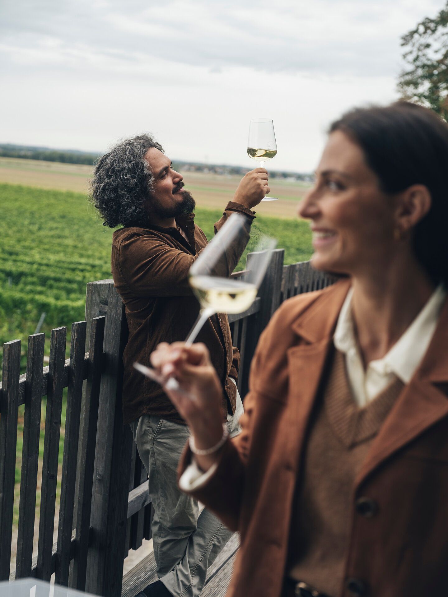 Zwei Personen stehen mit Weingläsern auf einer Terrasse mit Blick auf die Weinberge. Die Aufnahme entstand beim Weingut und der Vinothek Dam in der Ahrenberger und Eichberger Kellergasse im Traisental, Niederösterreich.