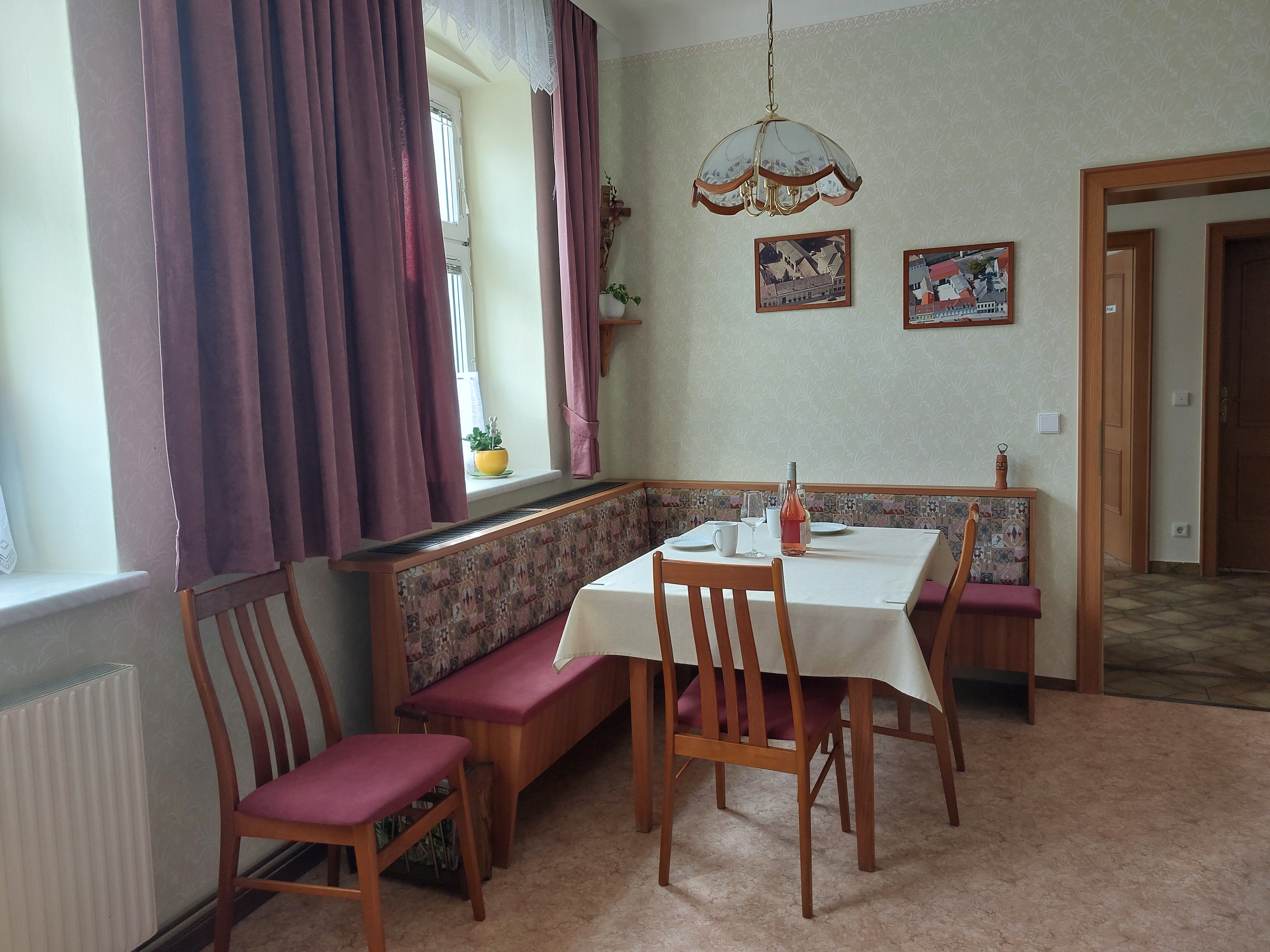 Hangulatos étkezős konyha étkezőasztallal, székekkel és sarokpaddal. Ablak függönyökkel, képek a falon.