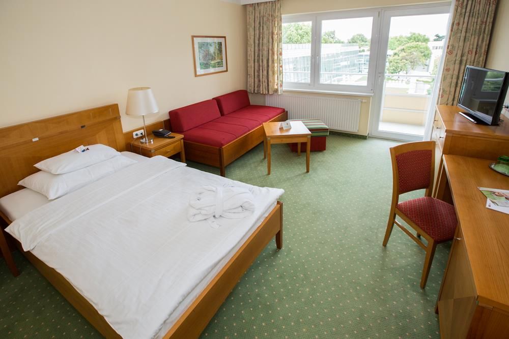 Hotelszoba franciaággyal, piros kanapéval, íróasztallal és erkéllyel.