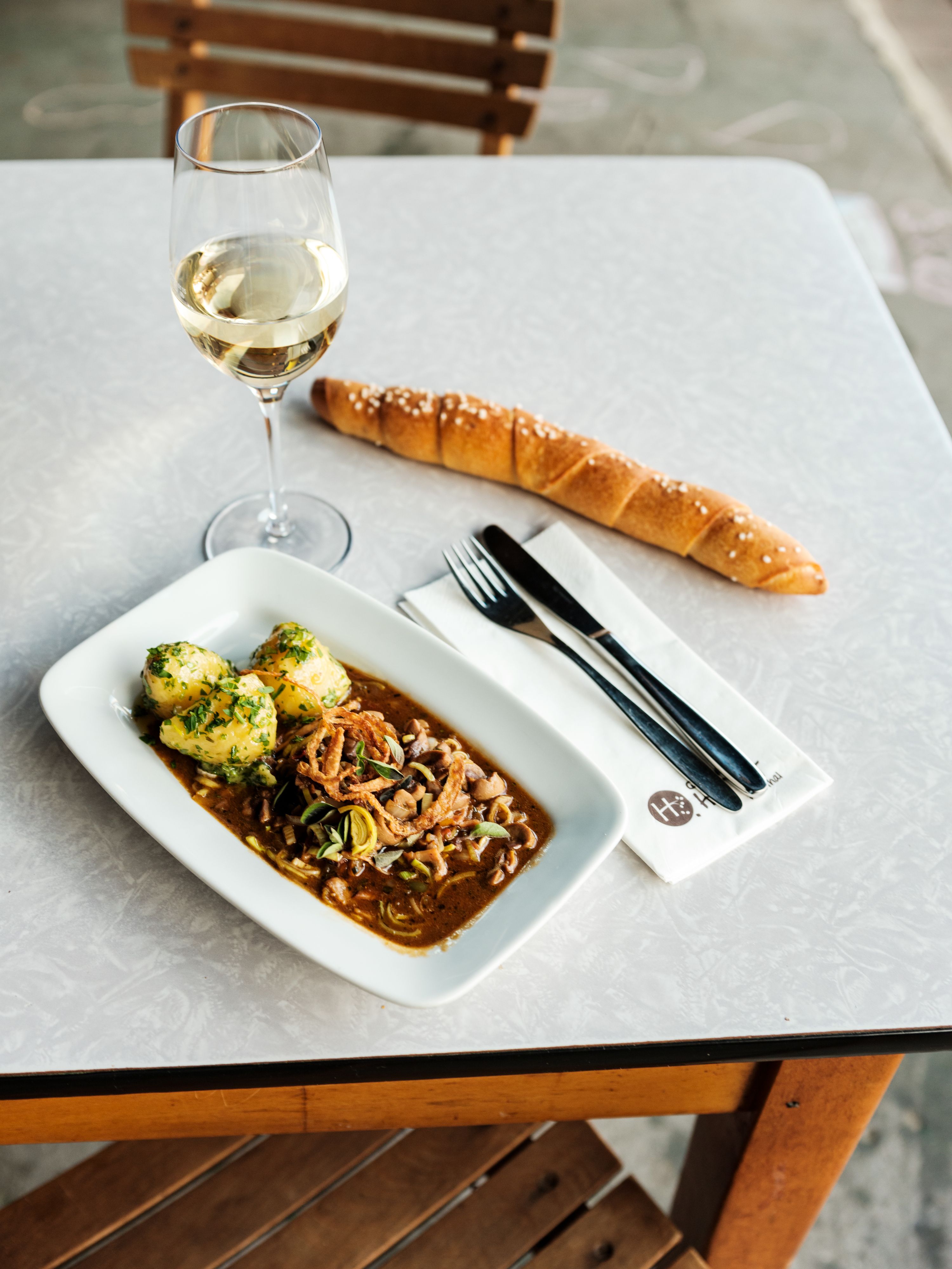 Egy asztal, rajta egy pohár fehérbor, egy tányér hústál és gombócok, egy hosszú zsemle és evőeszközök egy szalvétán.