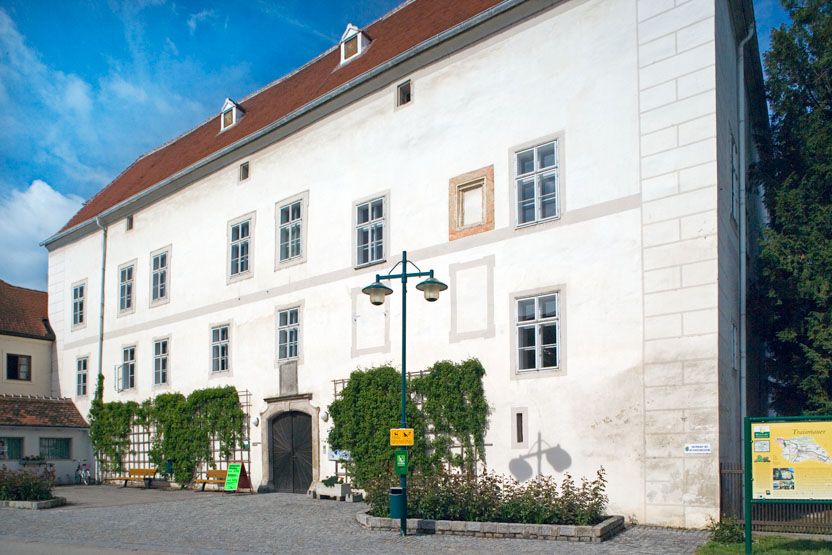 Traismauer kastély fehér homlokzattal és vörös tetőcseréppel, növényekkel és kövezett előkerttel körülvéve.