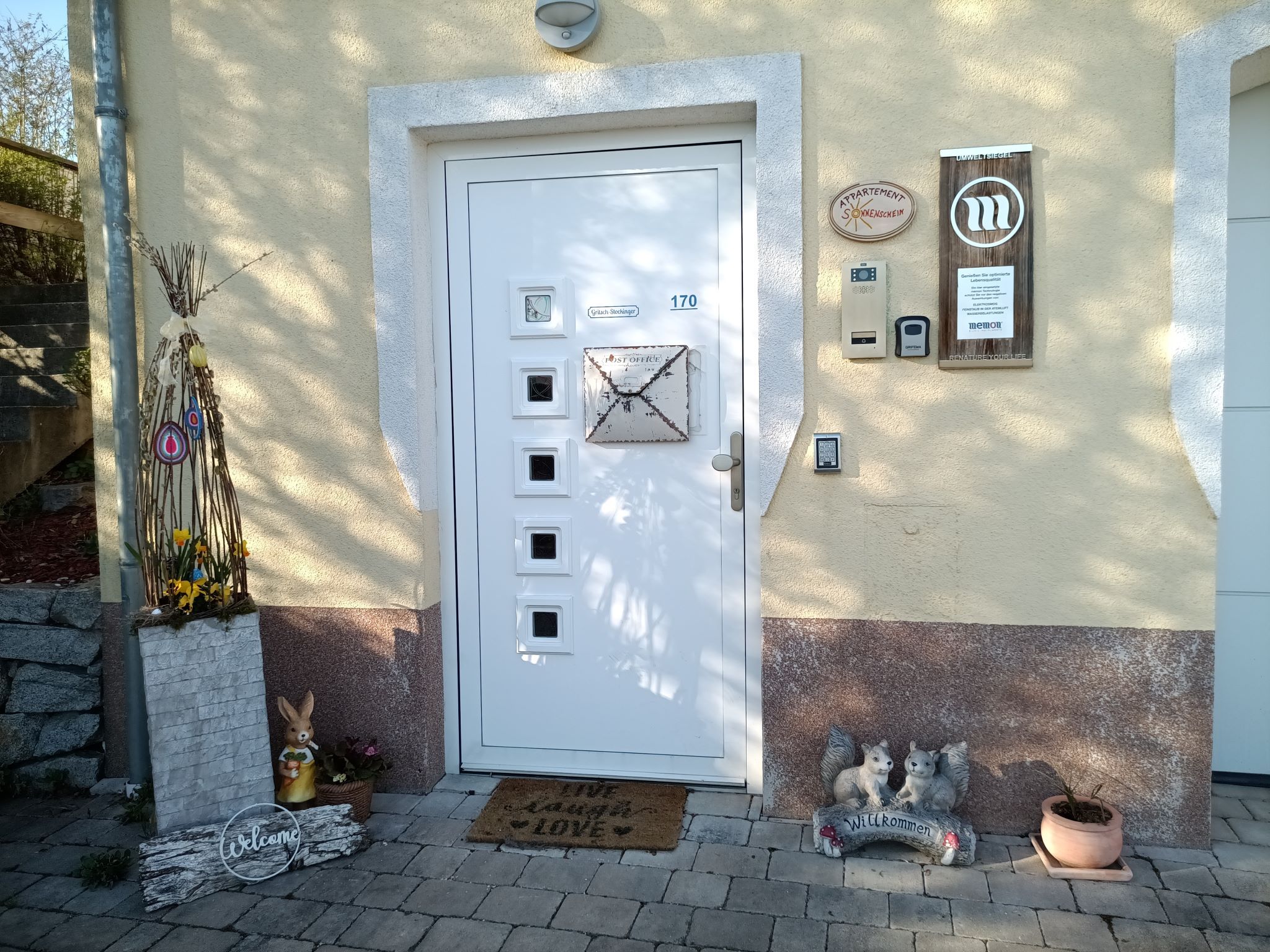 Egy ház bejárati része dekoratív elemekkel, például egy nyúllal, üdvözlő táblával és virágokkal.