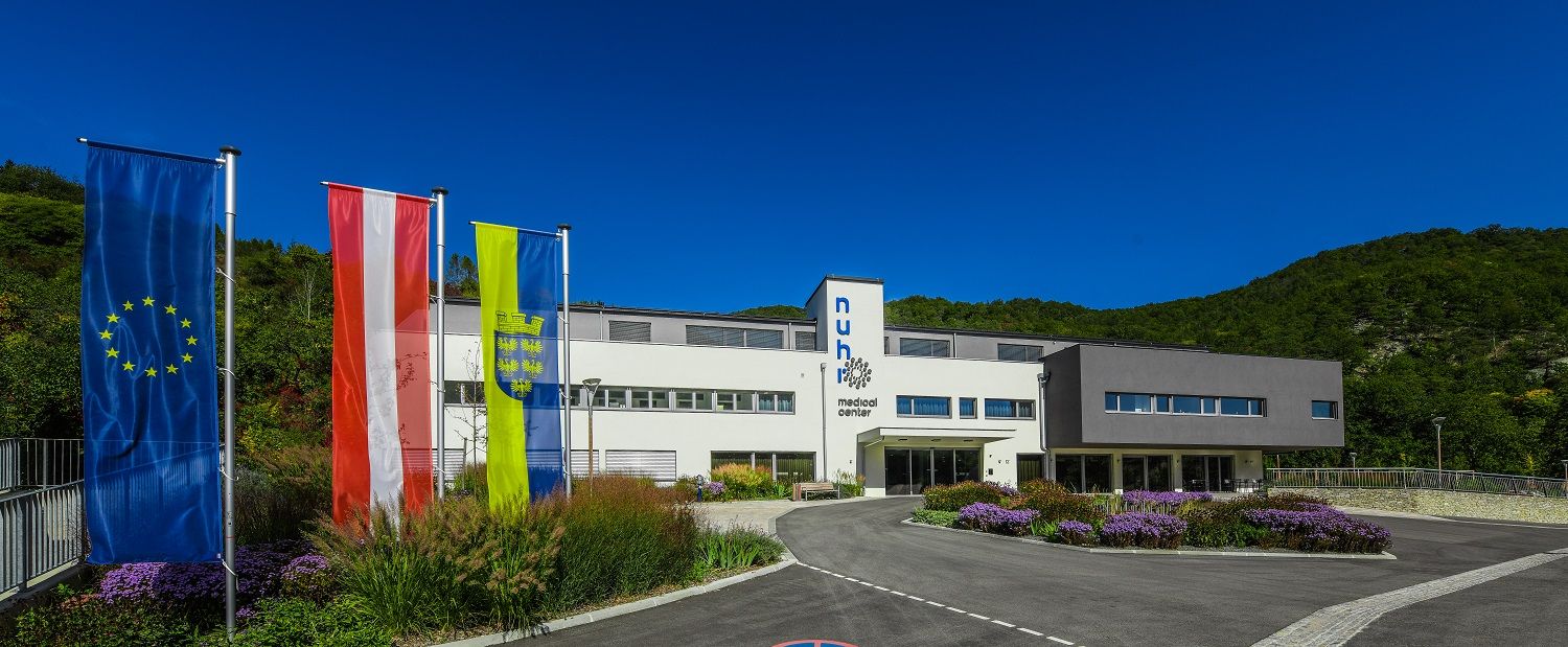 Modern épület három zászlóval az elején, zöld tájjal körülvéve.