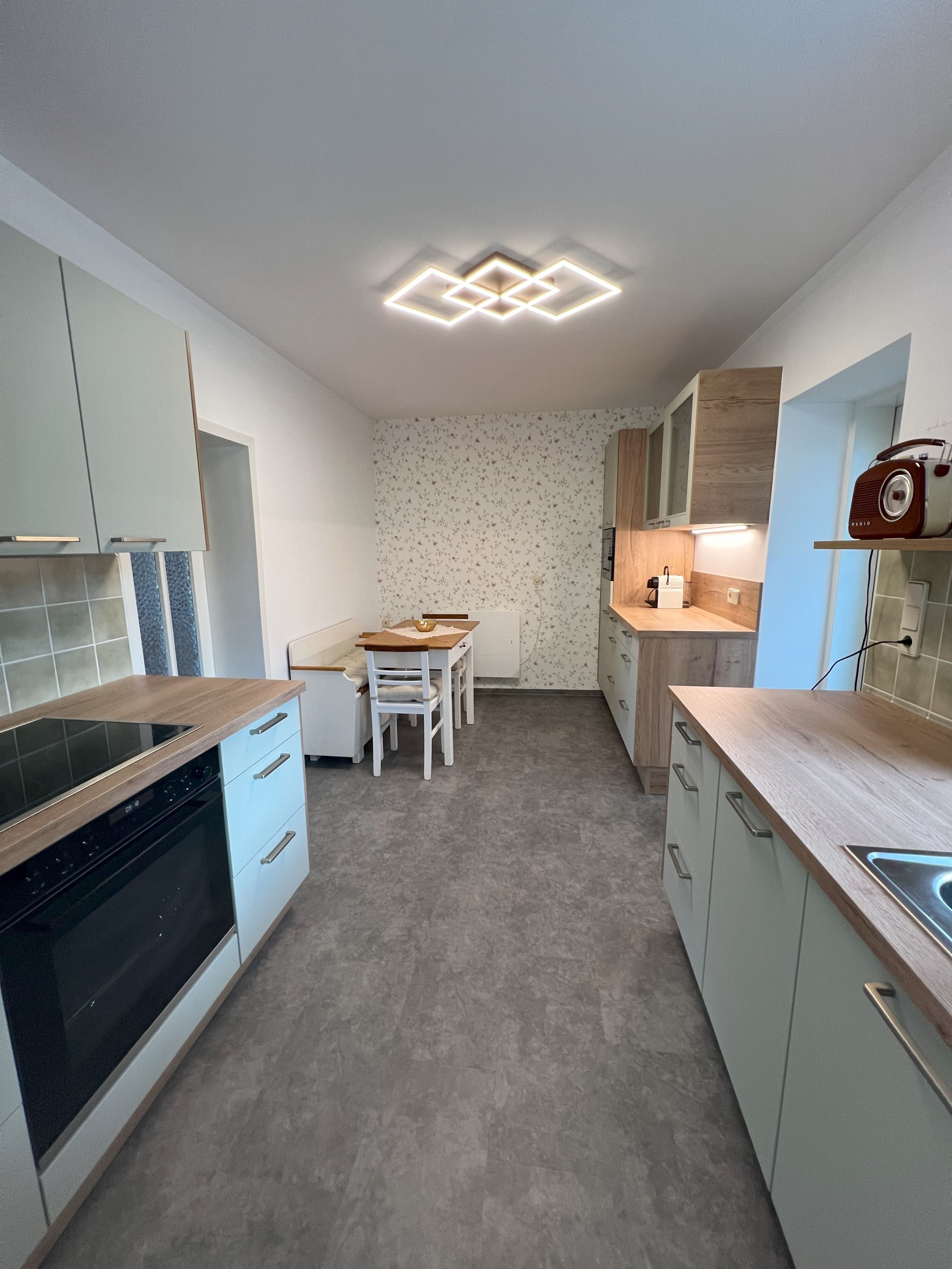 Modern konyha szürke szekrényekkel, fából készült díszítésekkel és étkezőasztallal. A mennyezeten geometrikus lámpa.