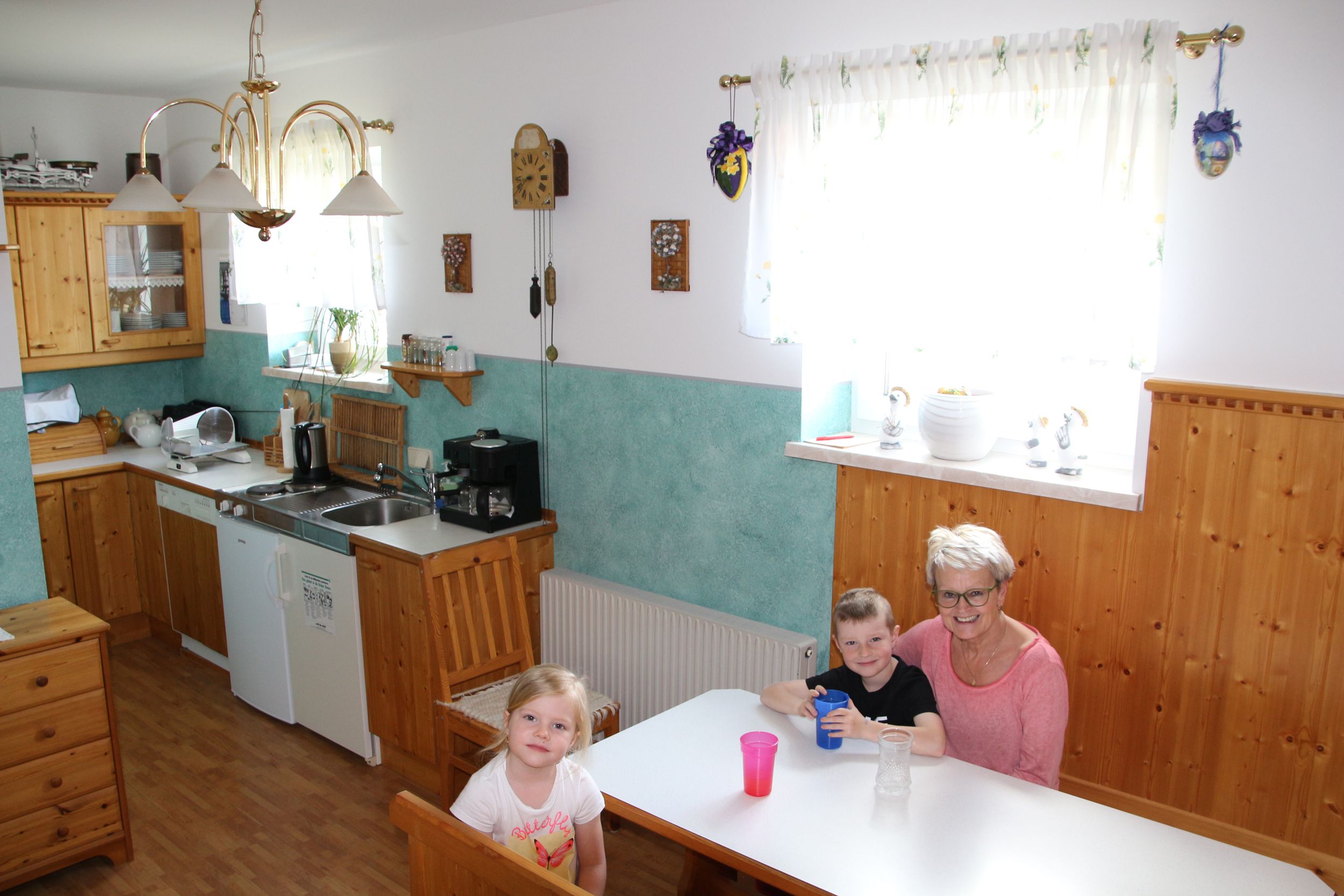 Egy reggelizőszoba fabútorokkal, a háttérben a konyha és három ember egy asztalnál.