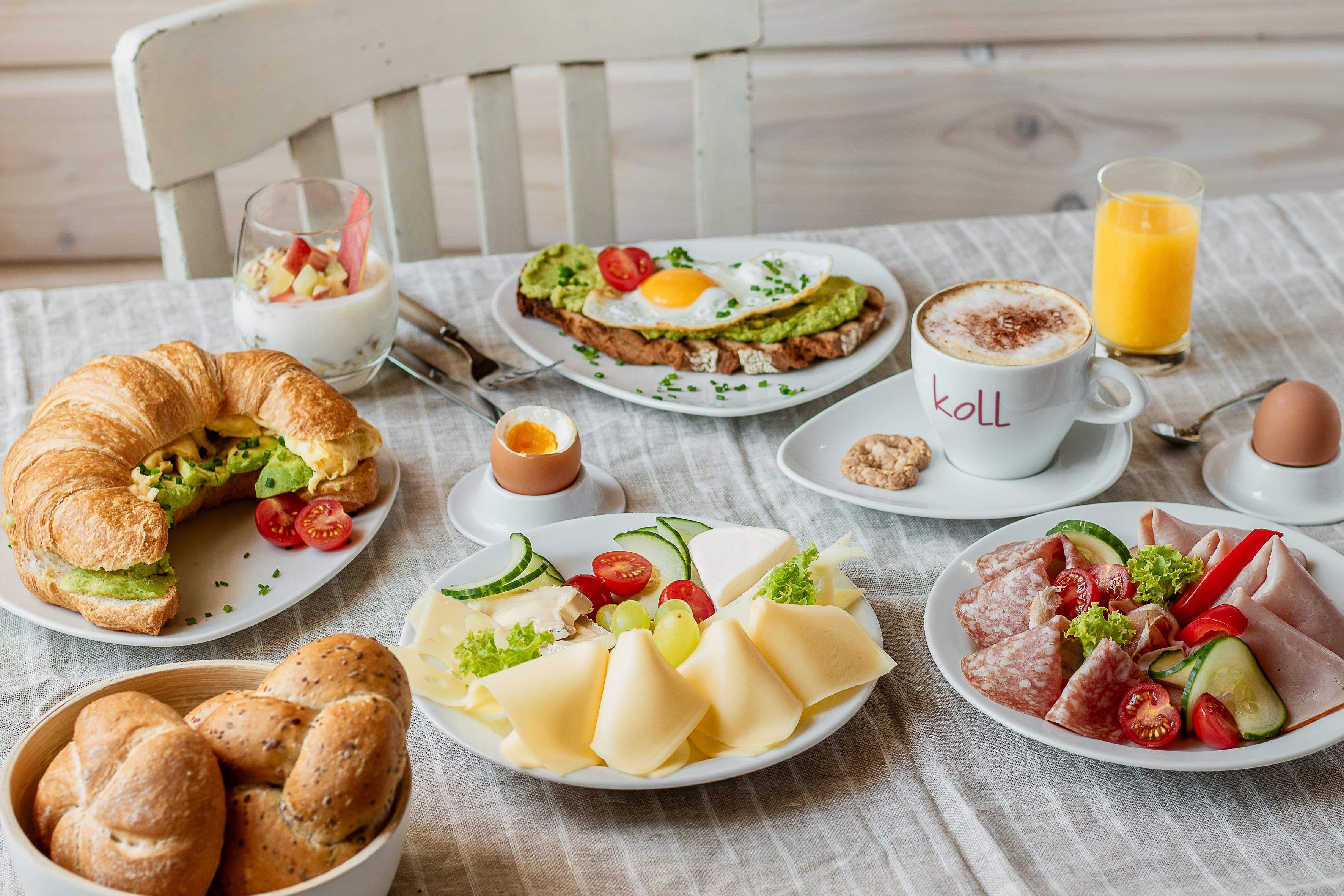 Bőséges reggeli croissant, kenyér, sajt, felvágott, tojás, joghurt, kávé és narancslé az asztalon.