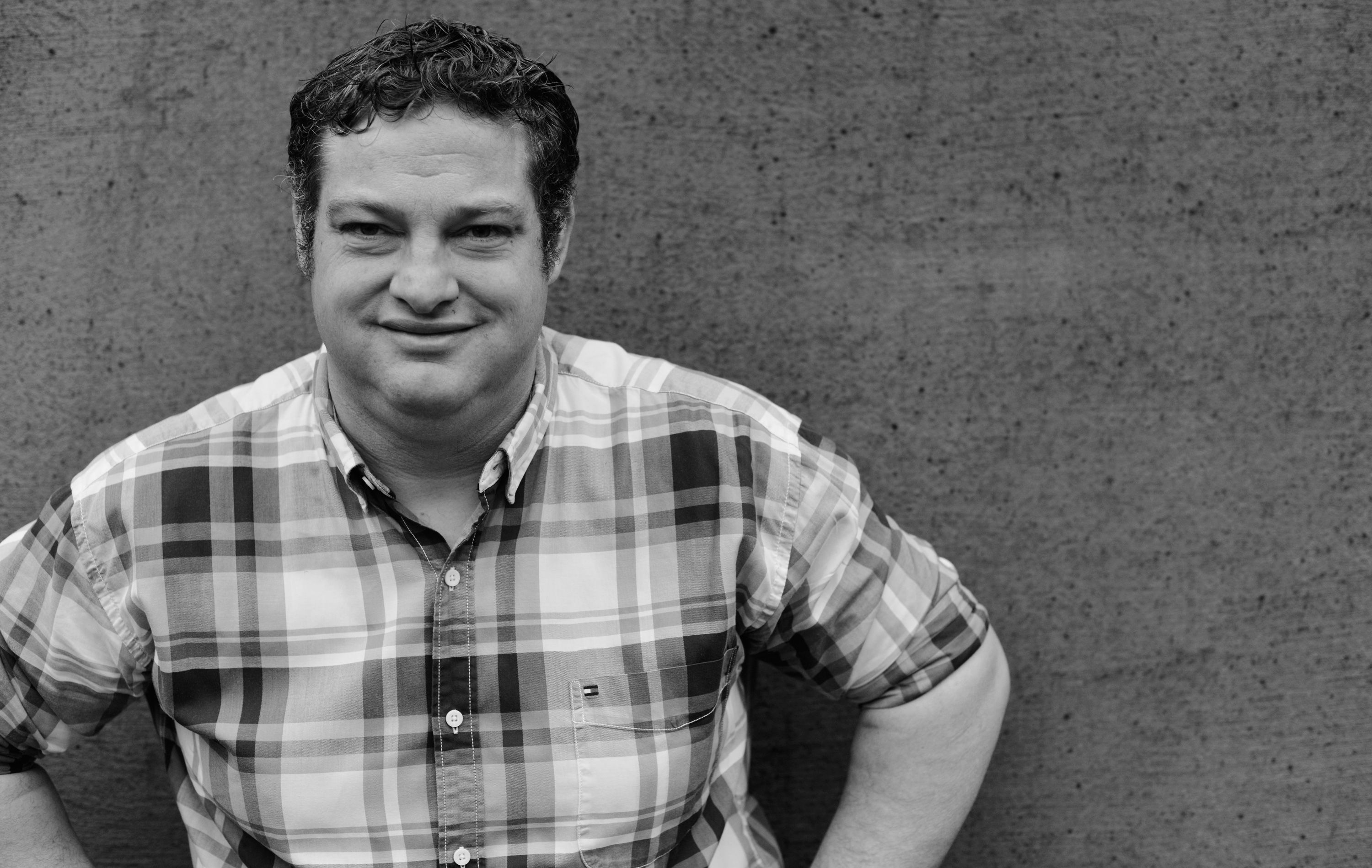 Fekete-fehér fotó egy kockás inges férfiról egy betonfal előtt.