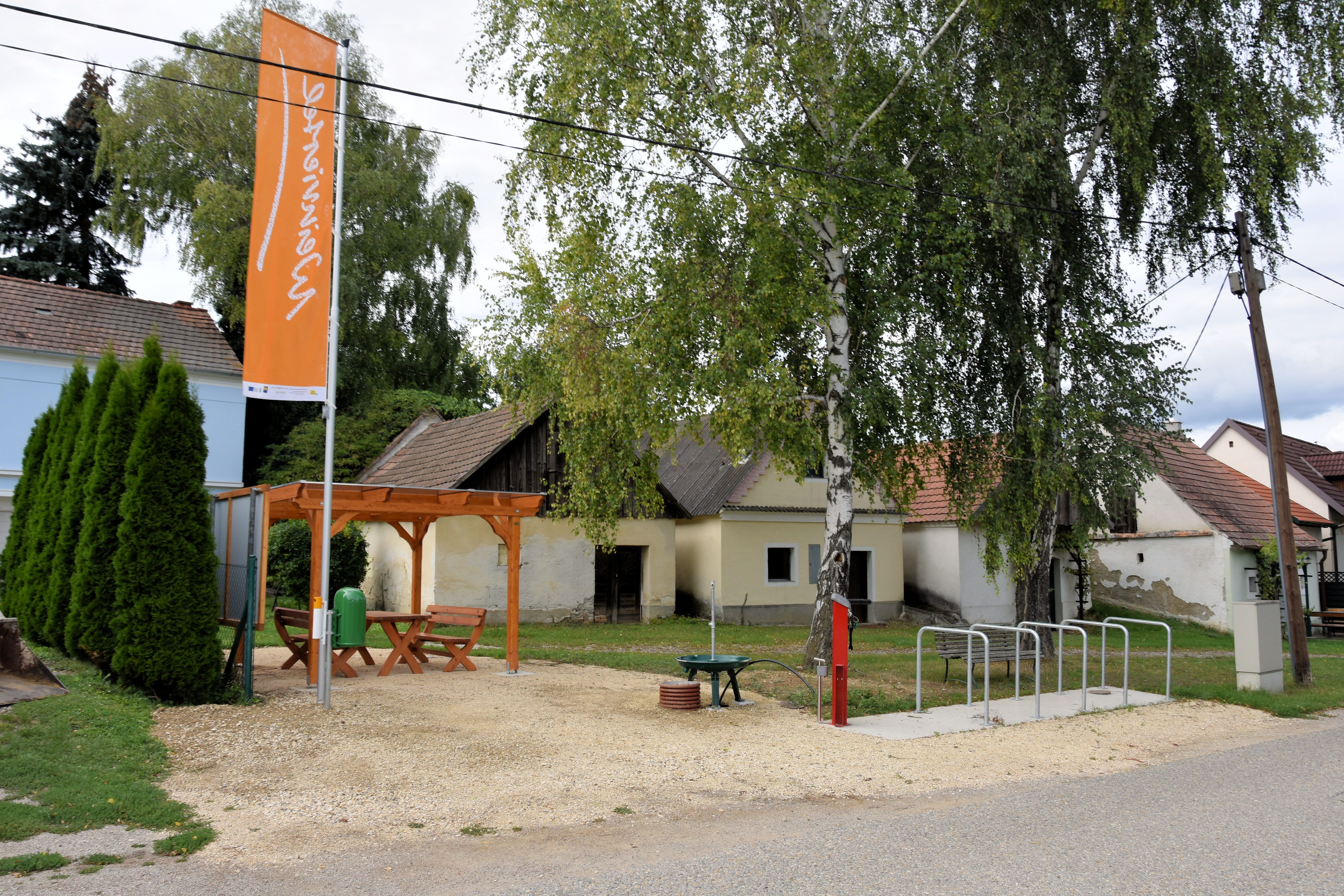 Egy pihenőhely, ahol egy pavilon alatt faasztal és padok találhatók, fákkal és régi épületekkel körülvéve. Kerékpártámasz és információs tábla is rendelkezésre áll.