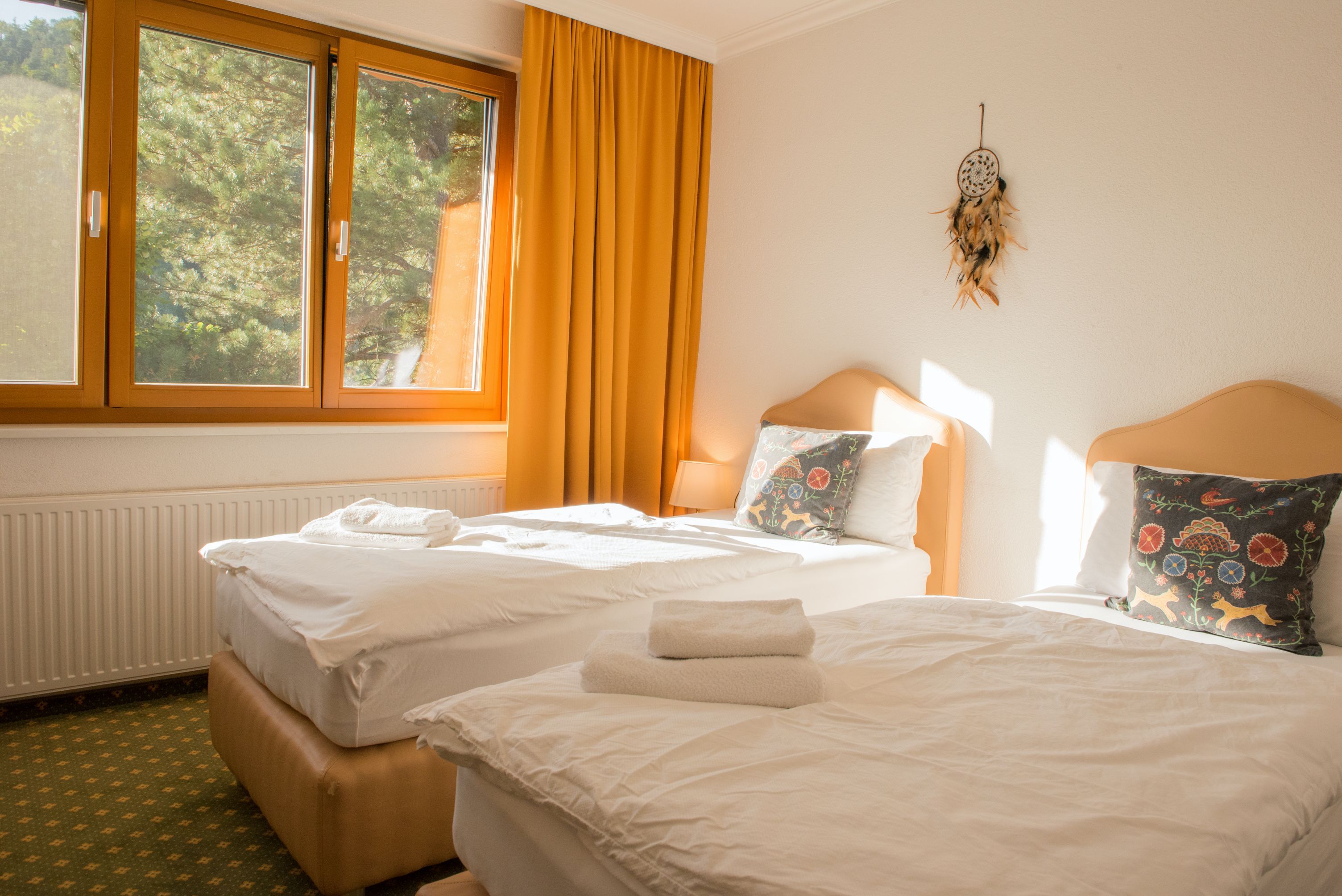 Világos kétágyas szoba két egyszemélyes ággyal, sárga függönyökkel és zöldre néző ablakkal.