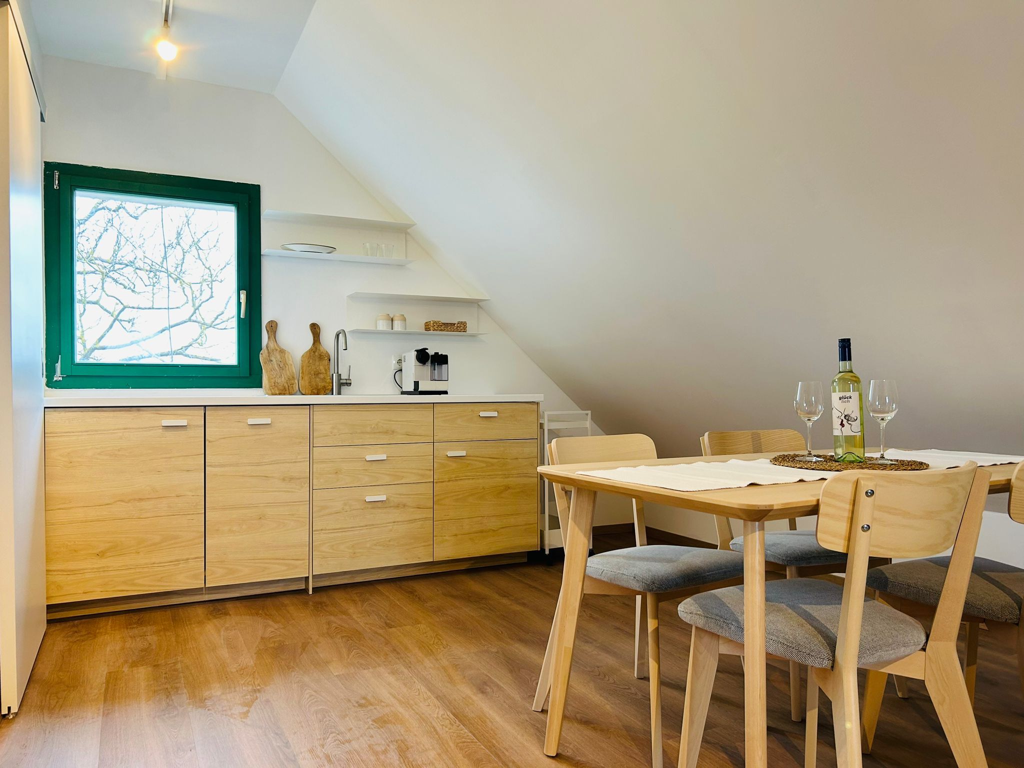 Modern konyha fa szekrényekkel, étkezőasztal és székek, zöld keretes ablak.