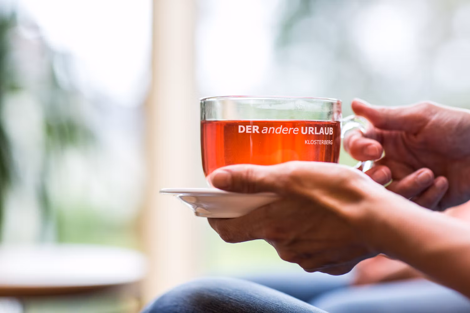 Közelkép a kezében piros itallal teli csészét tartó kezekről "DER andere URLAUB KLOSTERBERG" felirattal.