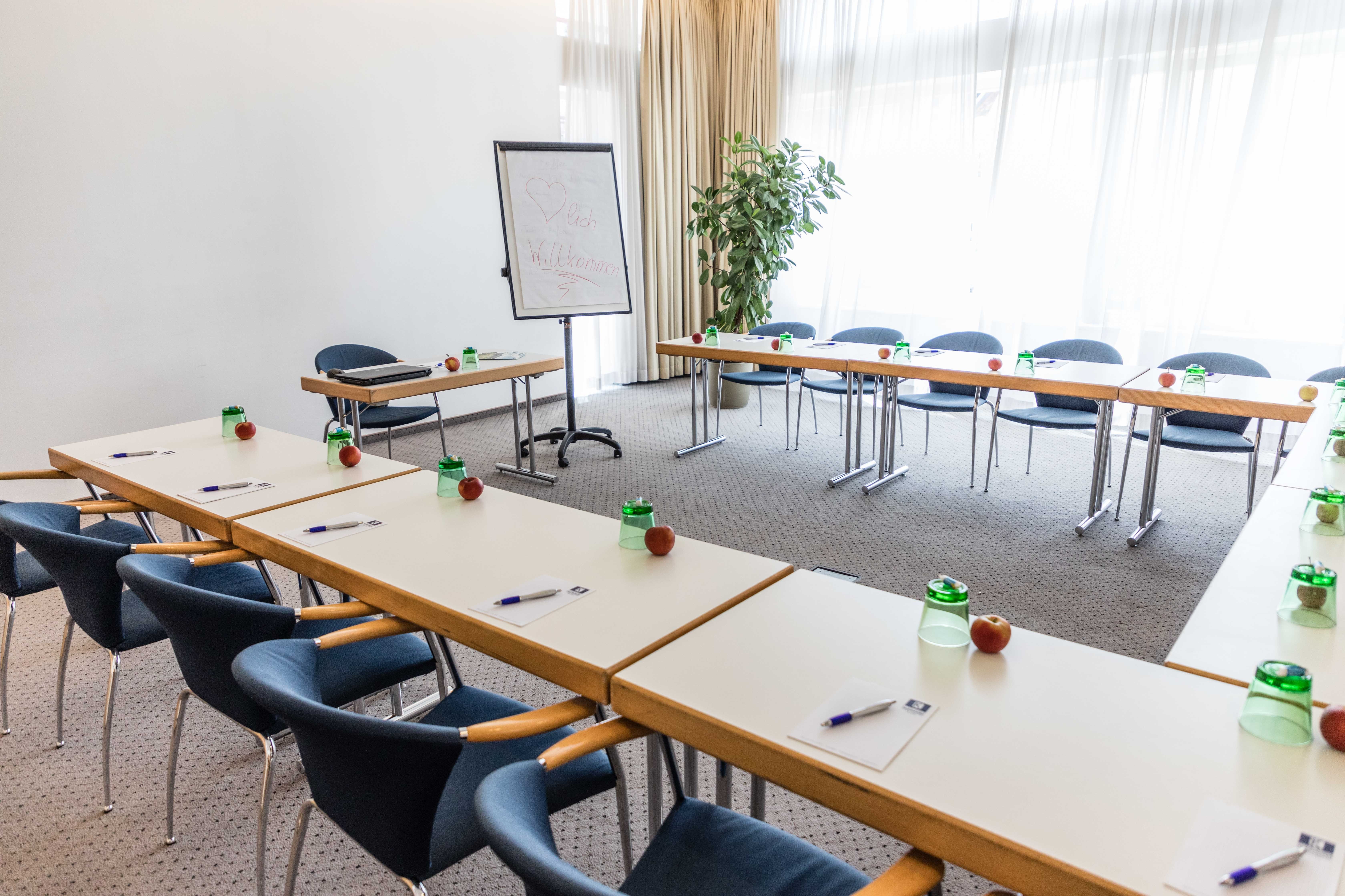 Konferenciaterem U alakú asztalokkal, székekkel, flipcharttal és íróeszközökkel.