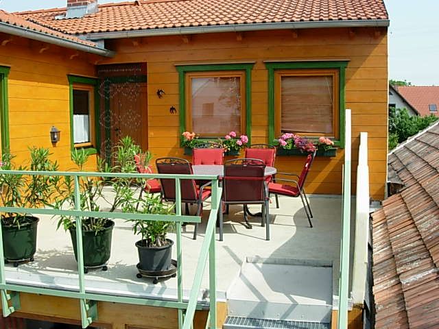 Balkon asztallal, székekkel és növényekkel, körülötte egy narancssárga színű homlokzatú ház, zöld ablakkeretekkel.