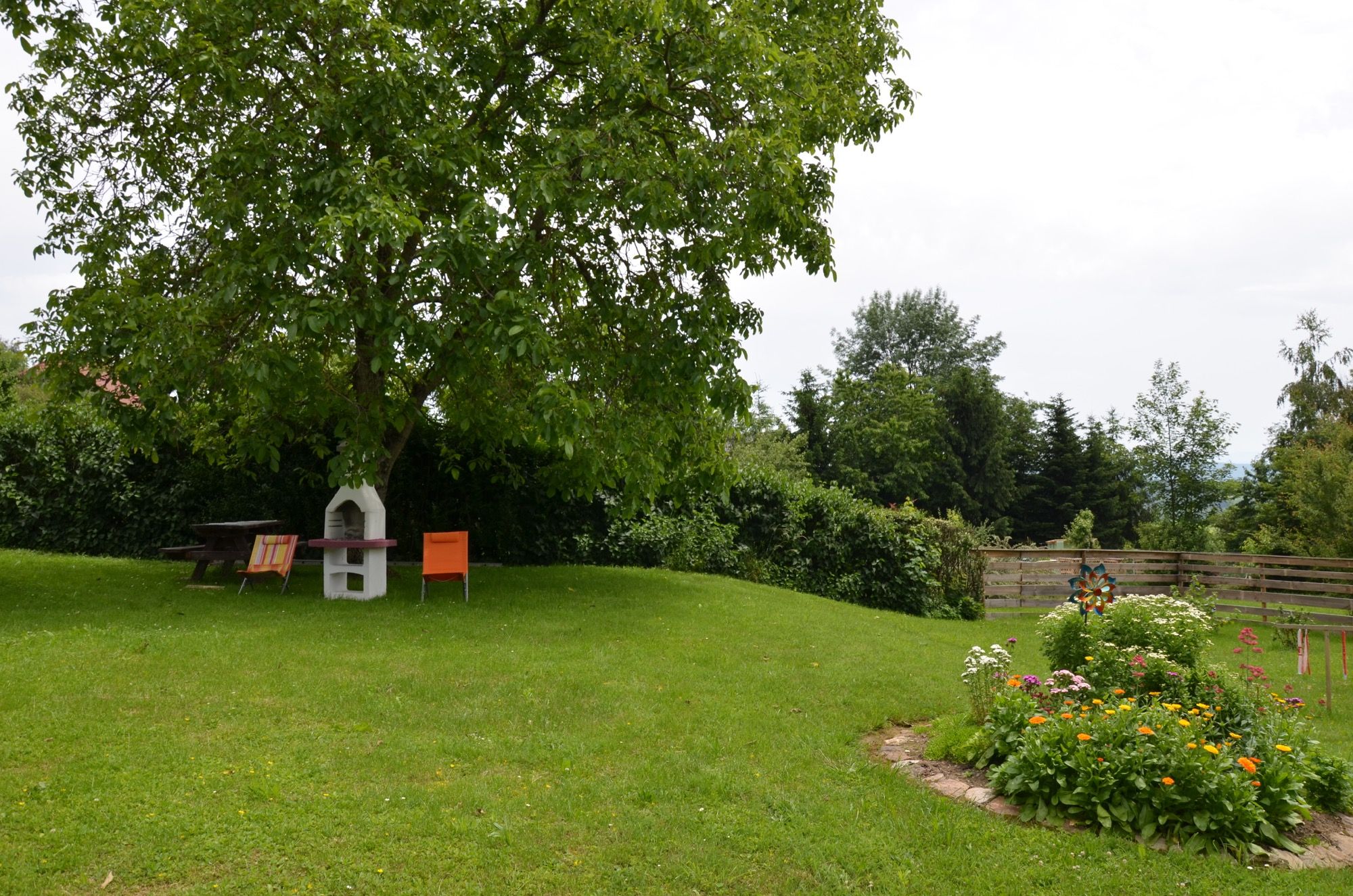 Zöld kert egy fával, kerti bútorokkal és virágágyással.