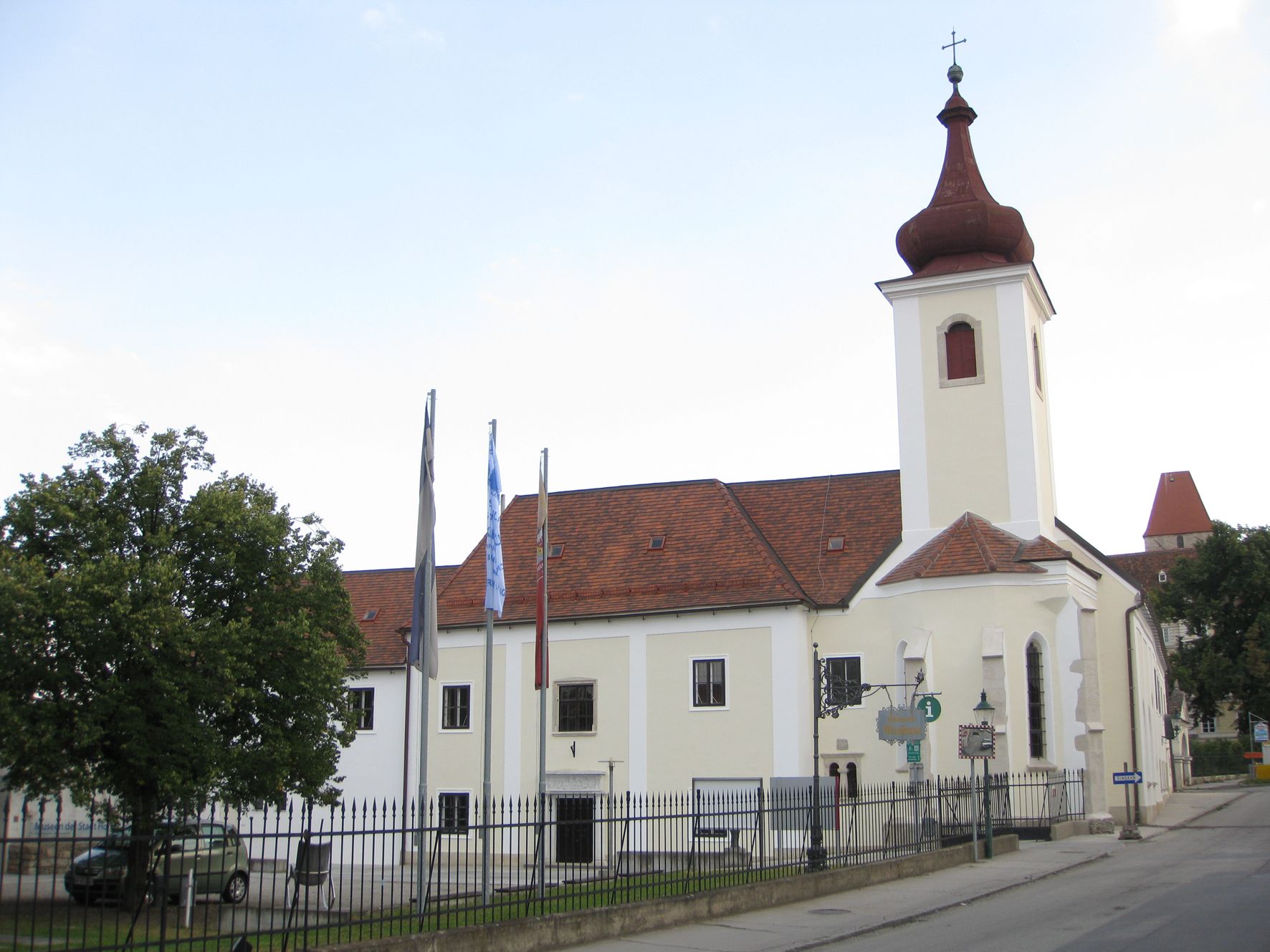 Egy templomtornyos, piros tetőcseréppel fedett épület, kerítéssel és fákkal körülvéve.
