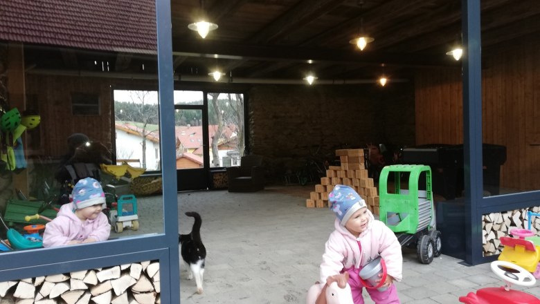 Egy téli ruhás gyermek játszik egy játéklovaggal egy nyitott szobában, ahol fakupacok és játékok vannak. Egy macska sétál el mellette.